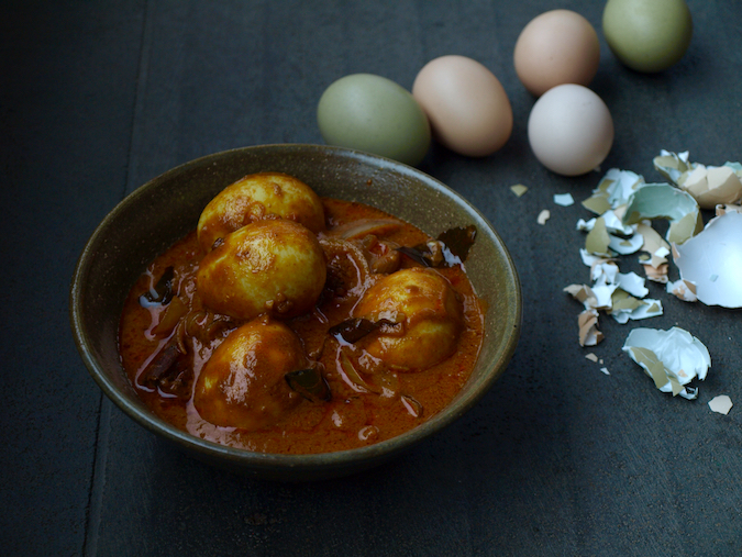 A bowl of egg curry. The eggs have been boiled in their shells, and the sauce is brick red with onions and curry leaves. Surrounding the plate are different coloured eggs and egg shells.
