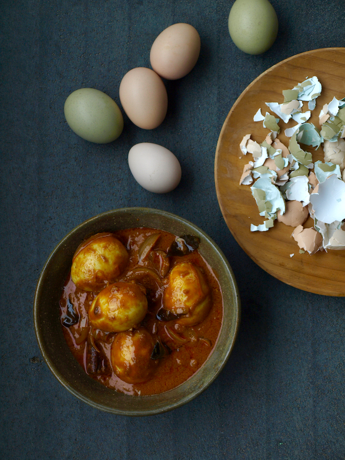 A bowl of egg curry. The eggs have been boiled in their shells, and the sauce is brick red with onions and curry leaves. Surrounding the plate are different coloured eggs and egg shells.