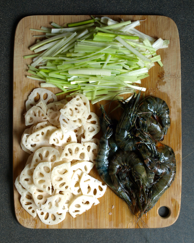 Raw tiger prawns (shrimp), julienned leek and sliced lotus root on a chopping board