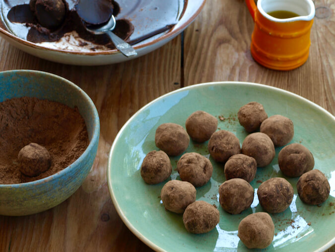 Olive oil chocolate truffles rolled in raw cacao powder on a plate. The bowl of mixture is in the background and an orange jug of olive oil.