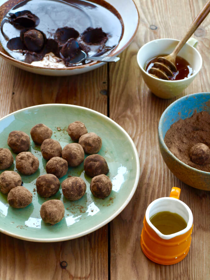 Olive oil chocolate truffles rolled in raw cacao powder on a green plate. In the photo there is also an orange jug of olive oil, a teacup of honey, and the bowl of truffle mixture in the background.
