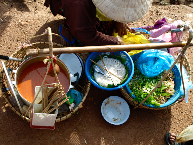 Roadside seller in southern Laos with a pot of brick red soup, fresh rice noodles, and salad