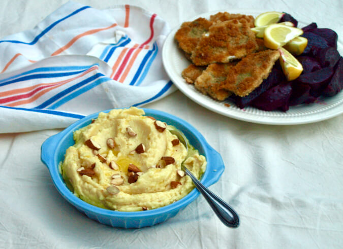 Bowl of Greek skordalia mayonaisa (mayonnaise) with almonds scattered on top. Plate of crumbed sardines, beetroot and lemon wedges in the background